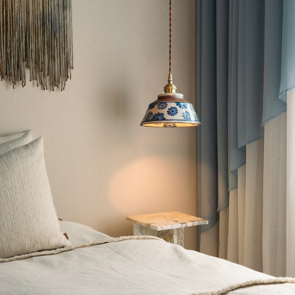 Lamp met blauwe bloemenkap hangt naast een bed met linnen kussens, een marmeren bijzettafel en blauwe gordijnen. 2