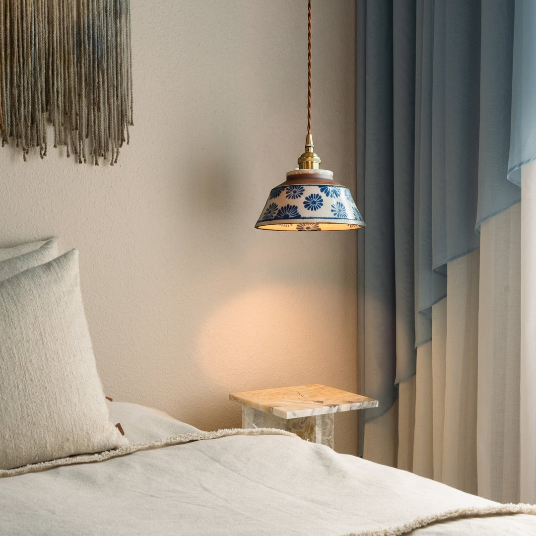 Lamp met blauwe bloemenkap hangt naast een bed met linnen kussens, een marmeren bijzettafel en blauwe gordijnen. 2