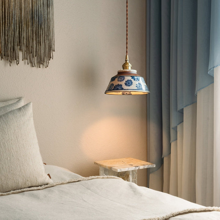 Lamp met blauwe bloemenkap hangt naast een bed met linnen kussens, een marmeren bijzettafel en blauwe gordijnen. 2