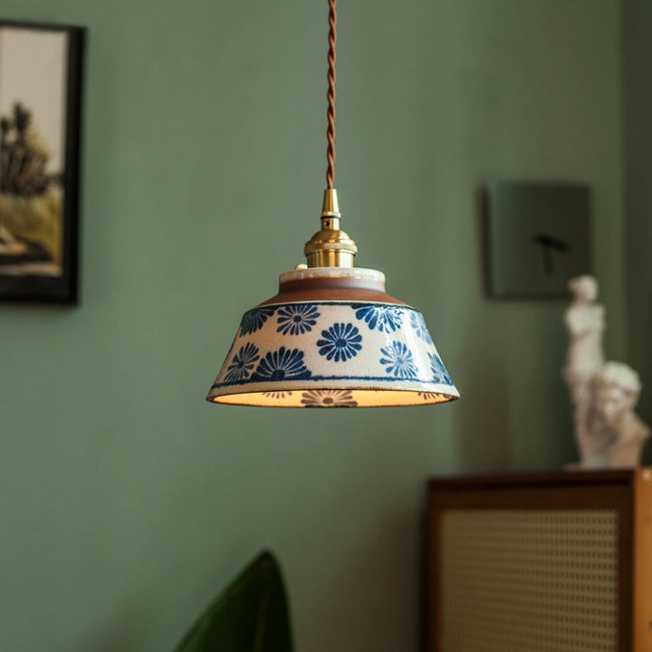 Brandende hanglamp met blauw bloemenpatroon op de keramische kap en gevlochten bruin snoer, voor een groene muur. 1