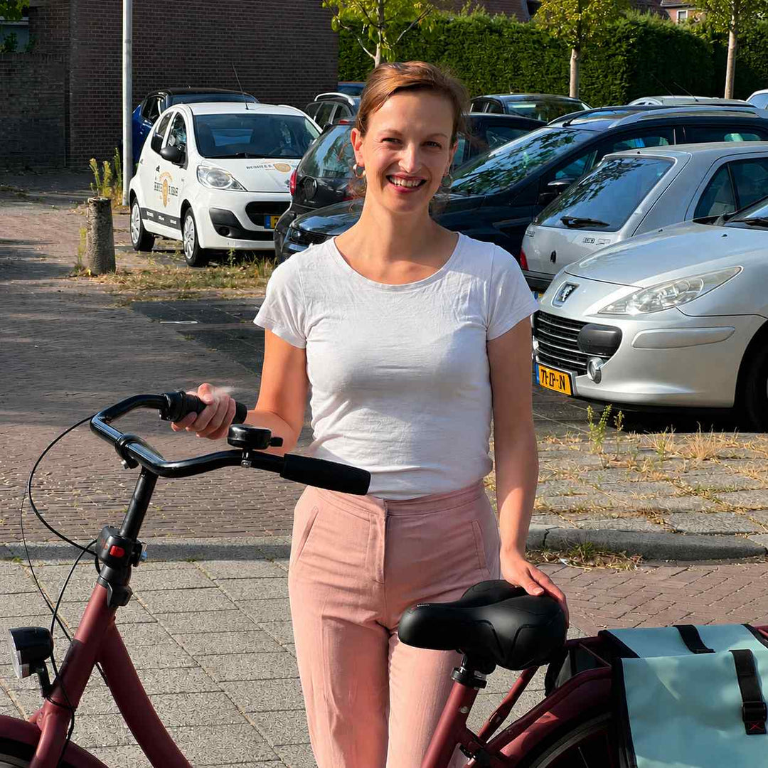 Lachende vrouw staat naast haar rode fiets en legt haar hand op het zwarte zadel, met fietstassen en geparkeerde auto's erachter. 7