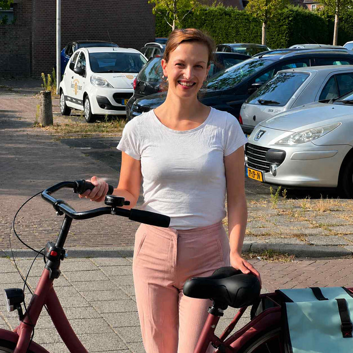 Lachende vrouw staat naast haar rode fiets en legt haar hand op het zwarte zadel, met fietstassen en geparkeerde auto's erachter. 7