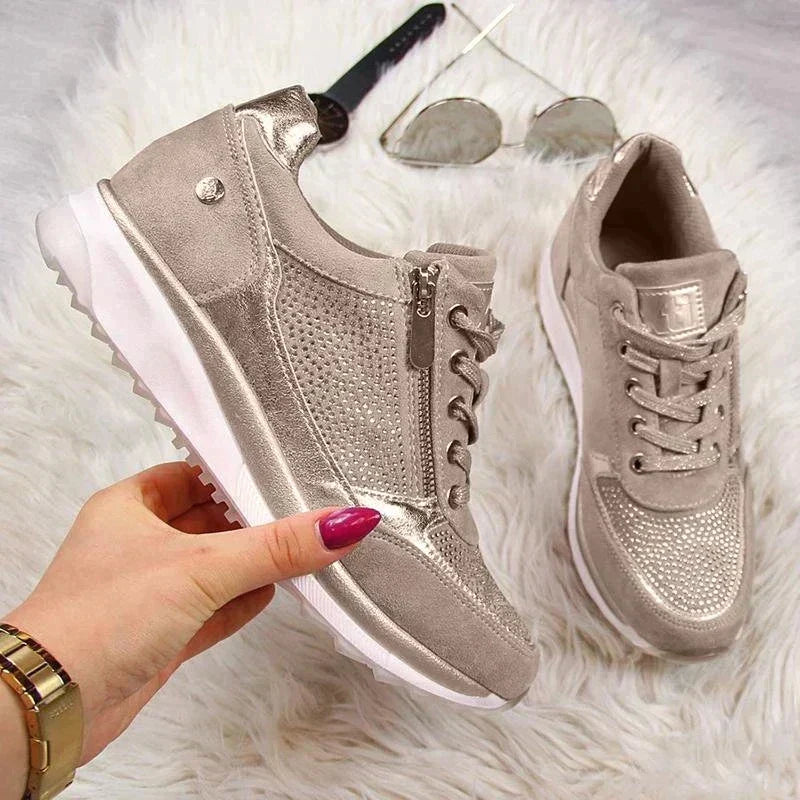 Twee taupe sneakers met zilverkleurige glans op een pluizig wit kleed, de linker schoen vastgehouden in een hand, naast zonnebril. 7