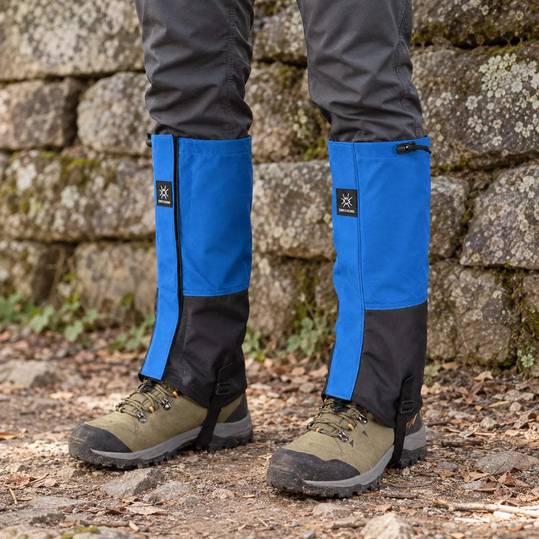 Blauwzwarte loopgamashen om wandelschoenen en broekspijpen, gedragen op een bospad voor een oude stenen muur met korstmos. 1