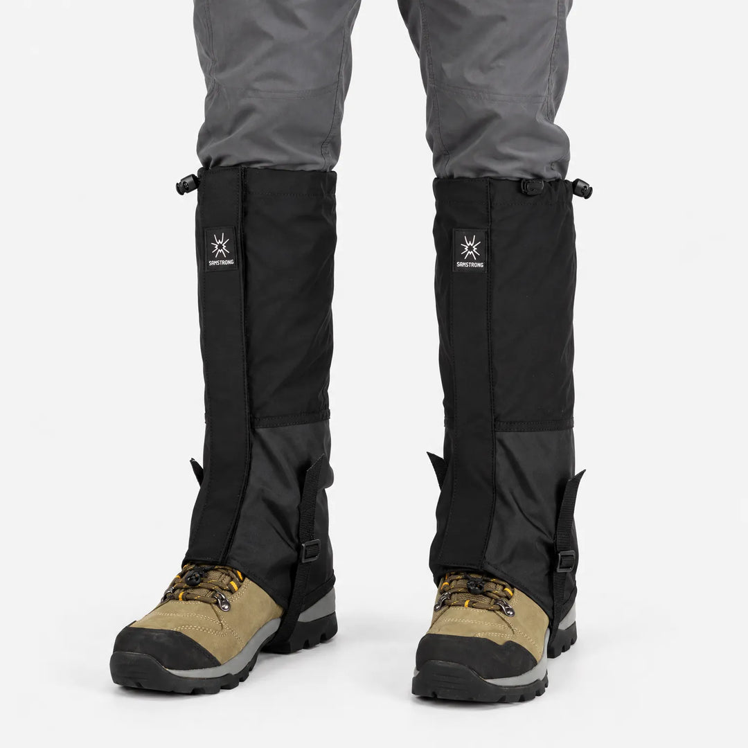 Zwarte loopgamashen gedragen boven bruine wandelschoenen en onder een grijze broek, frontaal gezien op een witte achtergrond. 8