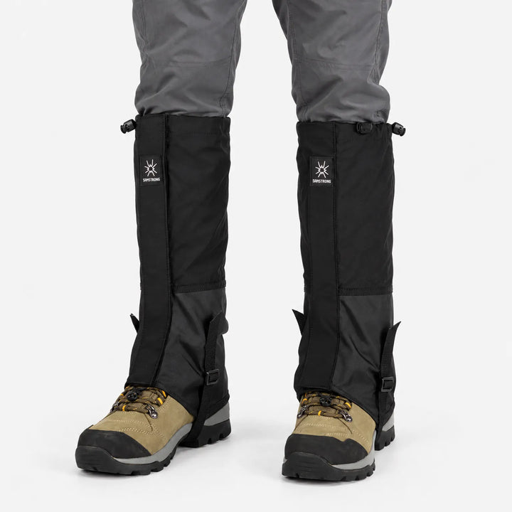 Zwarte loopgamashen gedragen boven bruine wandelschoenen en onder een grijze broek, frontaal gezien op een witte achtergrond. 8