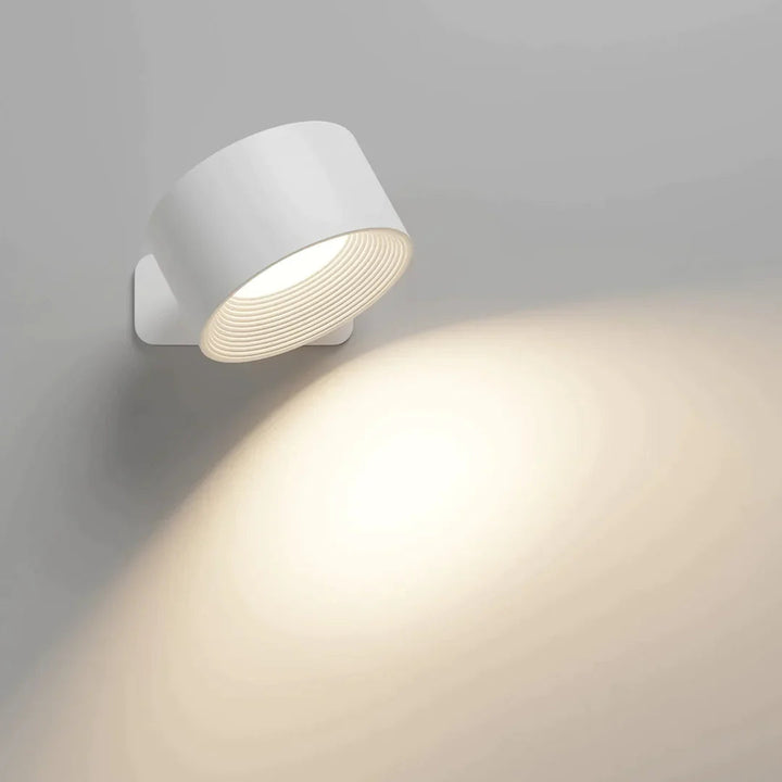 Witte ronde wandlamp met gegroefde binnenkant die een warme lichtbundel op een grijze muur werpt. 1