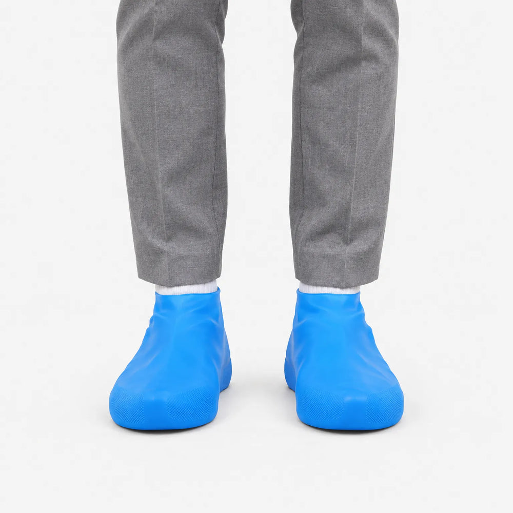 Voorkant van blauwe overschoenen aan de voeten van iemand in een grijze pantalon en witte sokken, op een witte achtergrond. 2