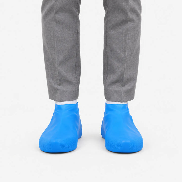 Voorkant van blauwe overschoenen aan de voeten van iemand in een grijze pantalon en witte sokken, op een witte achtergrond. 2