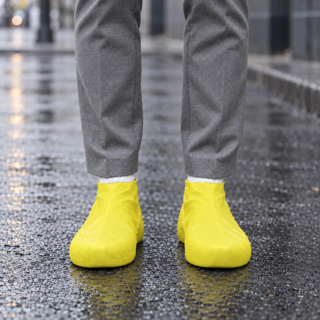 Gele overschoenen gedragen op een nat wegdek met waterdruppels erop, voorkant, bij iemand in een grijze pantalon. 3