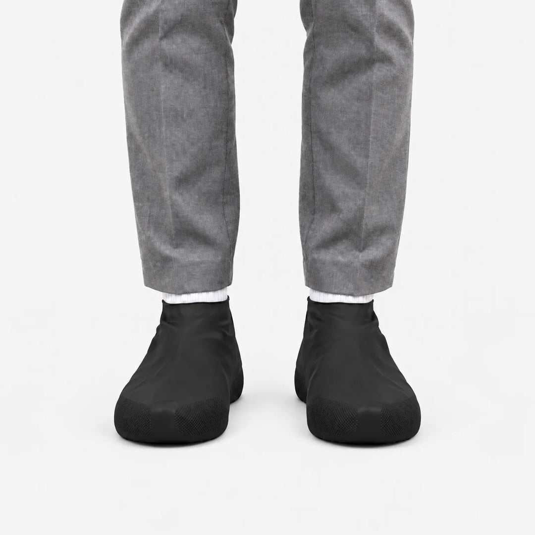 Zwarte overschoenen van voren gezien aan de voeten van iemand in een grijze broek, op een witte achtergrond. 4