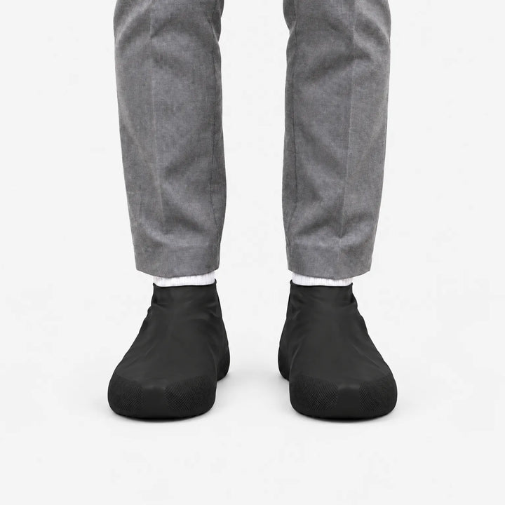 Zwarte overschoenen van voren gezien aan de voeten van iemand in een grijze broek, op een witte achtergrond. 4