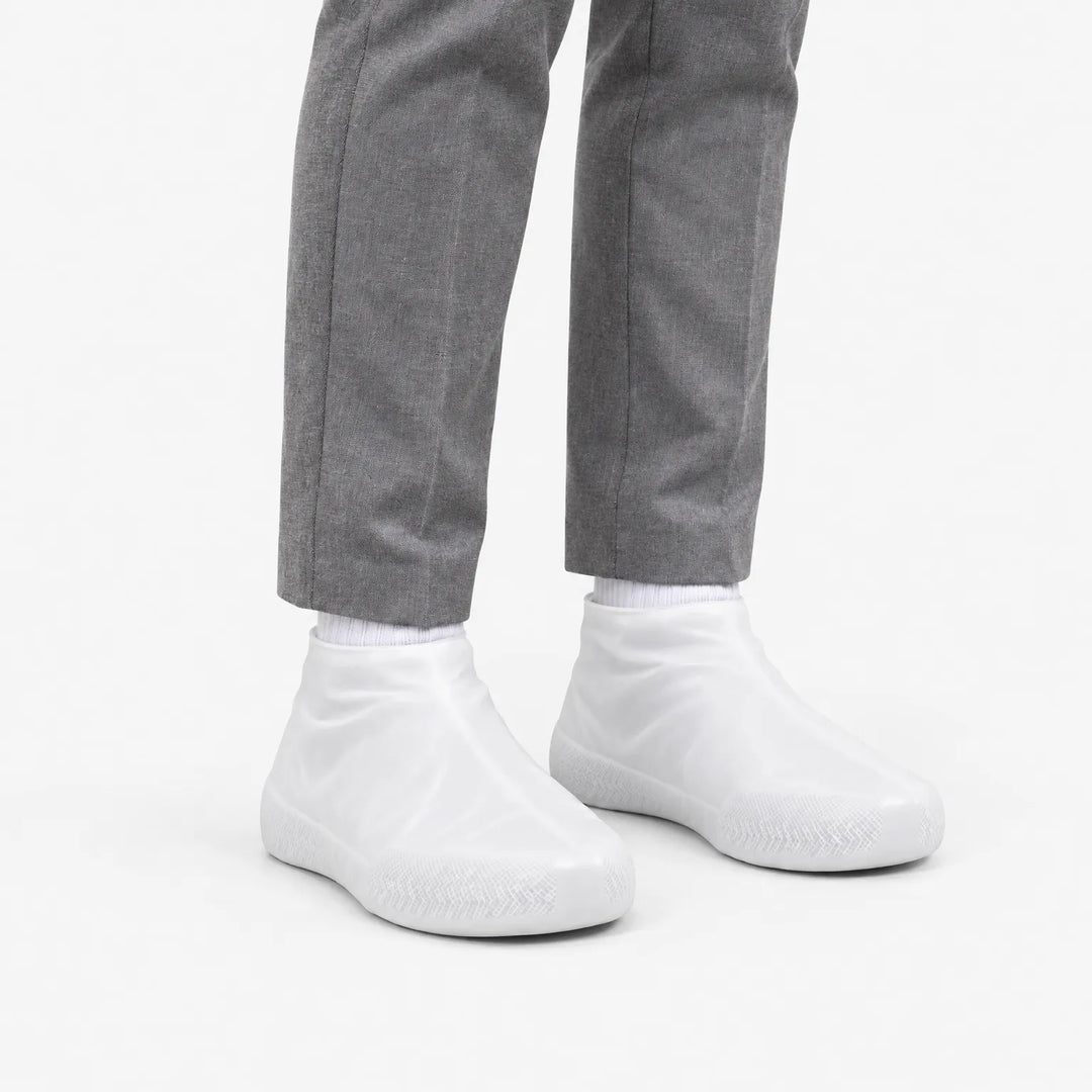Witte, doorschijnende overschoenen schuin van voren gezien, gedragen met een grijze pantalon en witte sokken. 5
