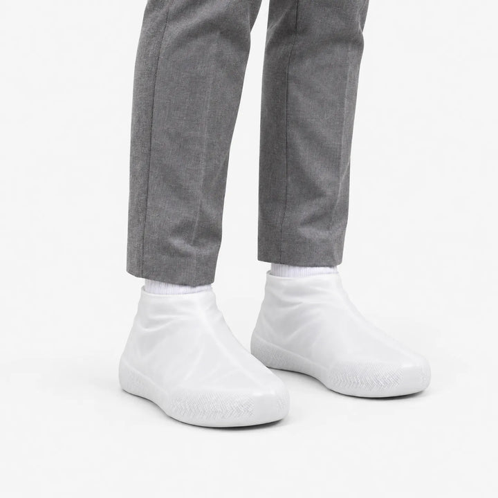 Witte, doorschijnende overschoenen schuin van voren gezien, gedragen met een grijze pantalon en witte sokken. 5