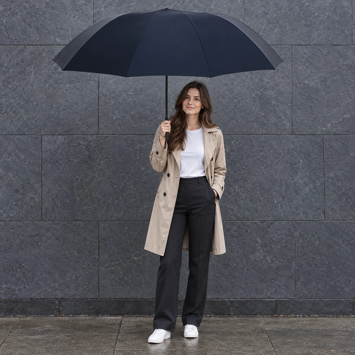 Vrouw in beige trenchcoat houdt een grote donkerblauwe paraplu boven zich tegen een grijze stenen muur op een natte stoep. 1