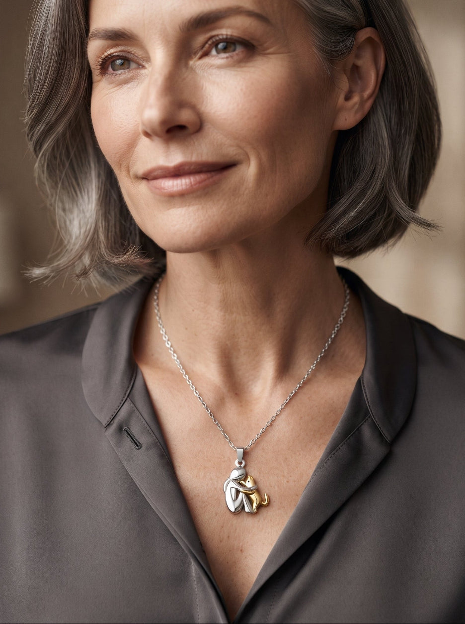 Vrouw met grijs bobkapsel en grijze blouse draagt de ketting met een zilverkleurige figuur die een goudkleurige huisdierfiguur omarmt. 10