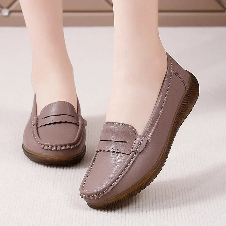 Taupekleurige instaploafers met lipje met sierrandje in plaats van gevlochten band, gedragen op een crèmekleurige vloer, zijaanzicht. 13