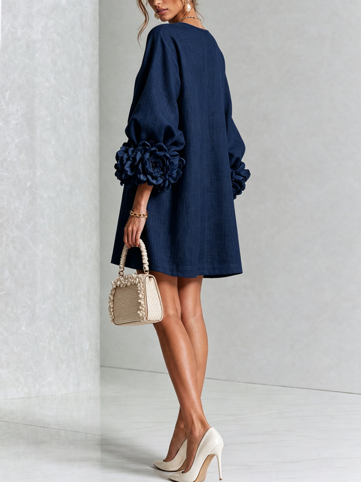 Model in de donkerblauwe jurk van achteren en schuin opzij, met roosmanchetten en een beige handtas met ruches. 13