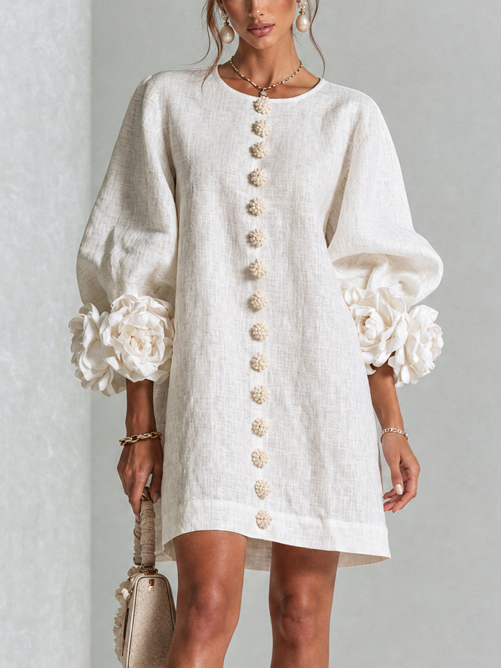 Model draagt de crèmewitte jurk van voren, met rij bloemknopen, pofmouwen en grote roosmanchetten, met een beige handtas in de hand. 1