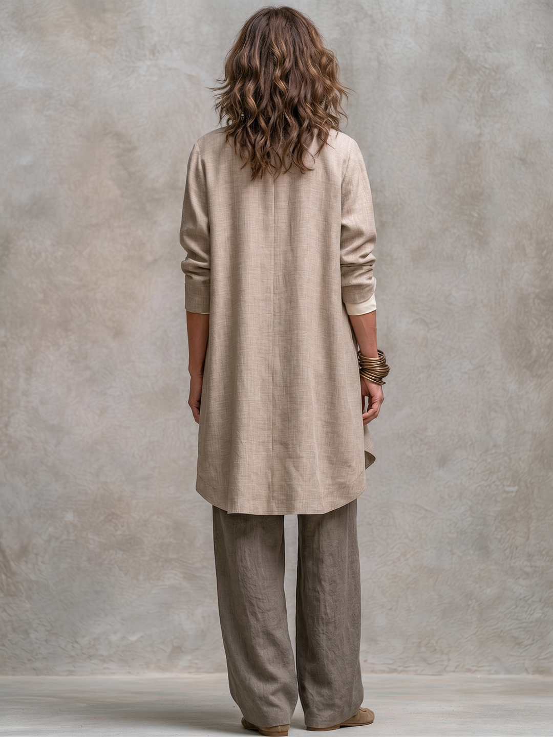Model van achteren: de lange beige jack met middennaad en ronde zoom, gedragen over de taupe broek. 6