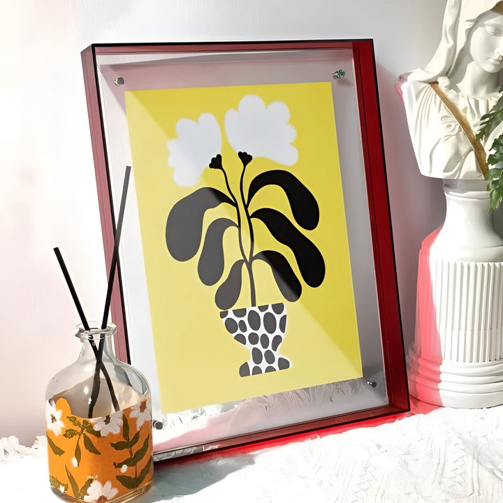 Rode acryllijst met gele bloemenprint, schuin tegen de muur geplaatst naast een geurstokjesflesje en een wit beeldje. 15