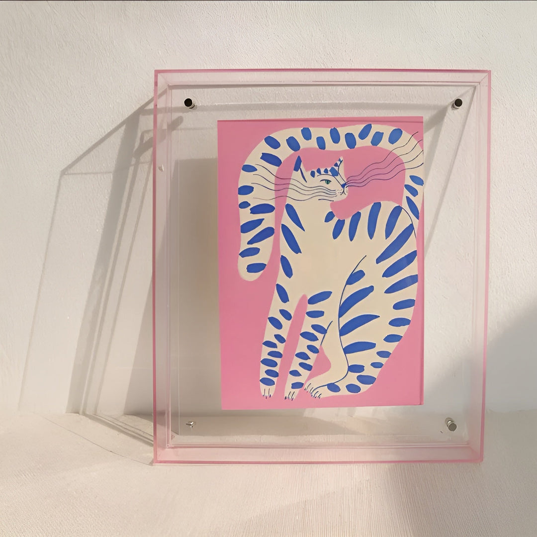 Transparante acryllijst met roze rand tegen een witte muur, gevuld met een roze illustratie van een wit-blauw gestreepte kat. 9