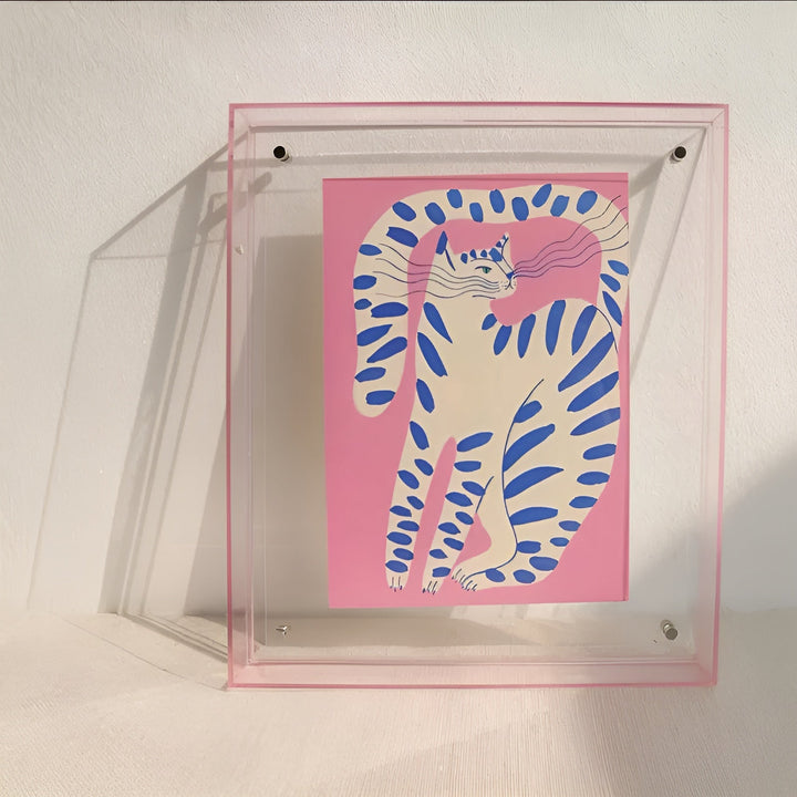 Transparante acryllijst met roze rand tegen een witte muur, gevuld met een roze illustratie van een wit-blauw gestreepte kat. 9