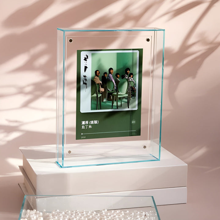 Transparante acrylfotolijst met blauwgroene randen staat op een witte doos, met een groen albumhoesje van een groep mensen op stoelen erin. 13