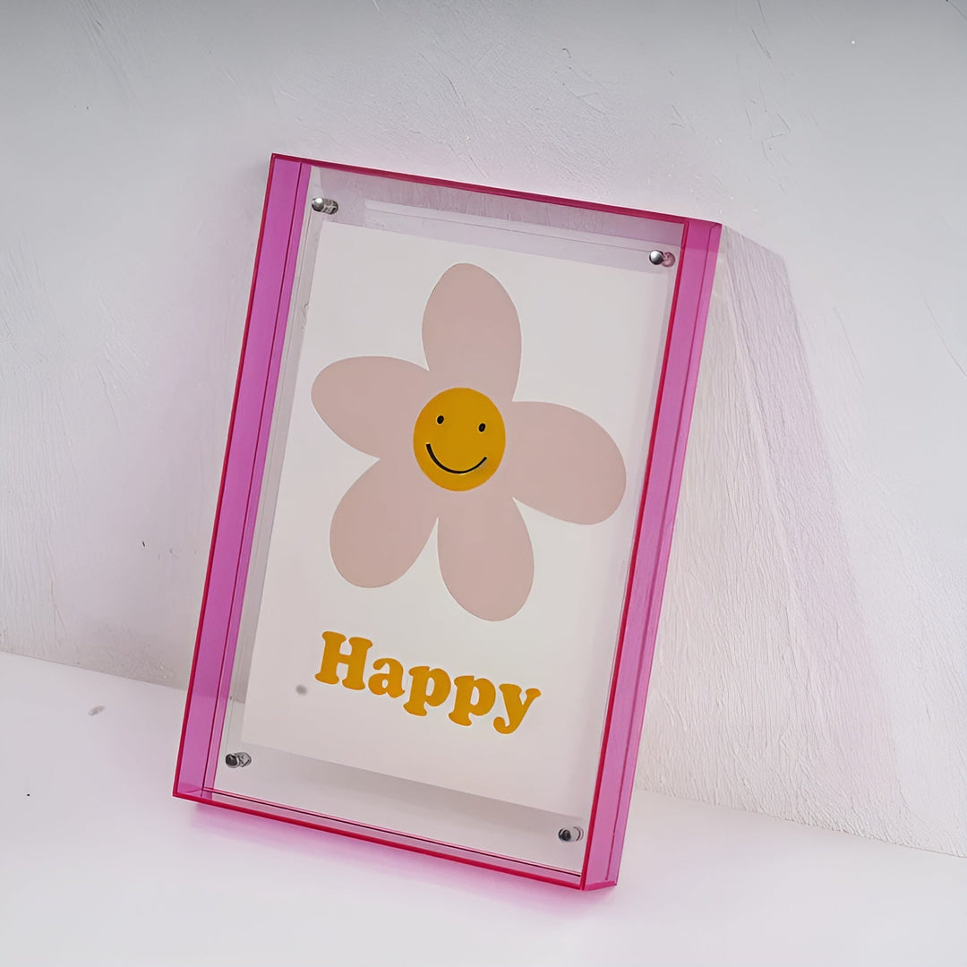 Roze acrylfotolijst met doorzichtige voorkant, schuin tegen een witte muur geplaatst, met een poster van een lachende bloem erin. 19
