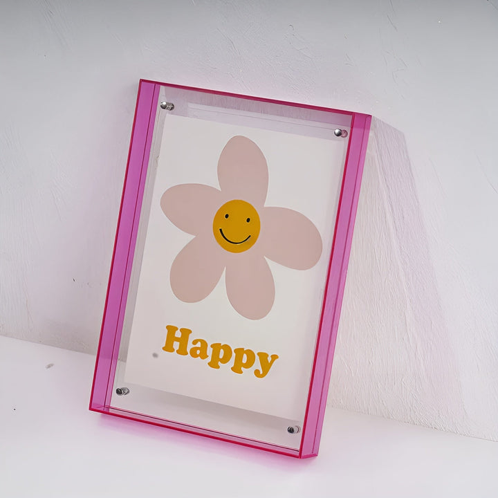 Roze acrylfotolijst met doorzichtige voorkant, schuin tegen een witte muur geplaatst, met een poster van een lachende bloem erin. 19