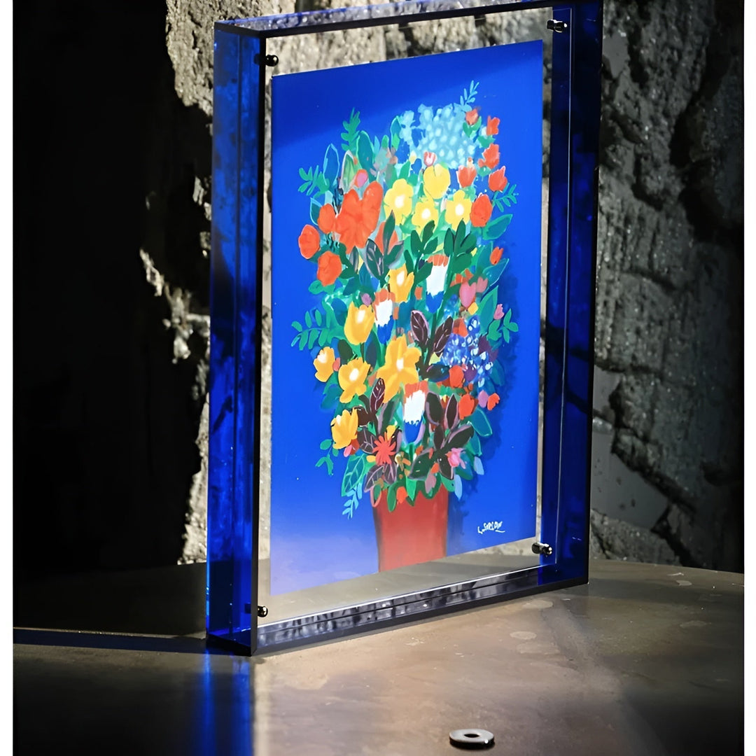 Blauwe acryl fotolijst met kleurrijk bloemenschilderij, schuin opgesteld op een tafel voor een ruwe stenen muur. 4