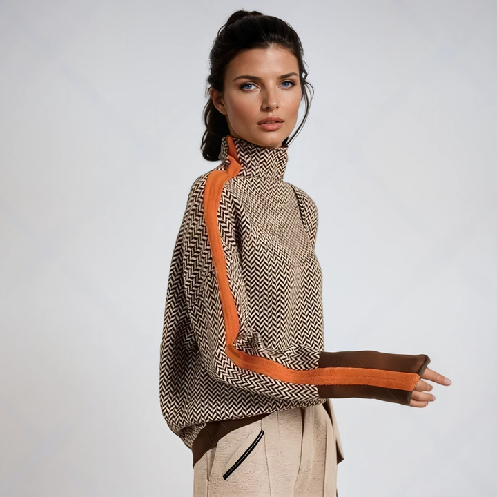 Vrouw in zijaanzicht draagt de trui met visgraatpatroon, oranje streep langs de mouw en bruine manchetten, gecombineerd met een beige broek. 1