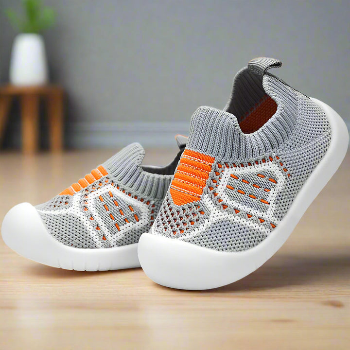 Paar grijze gebreide babyschoentjes met oranje streepdetails, ademend gaas en dikke witte zool, schuin geplaatst op een houten vloer. 7