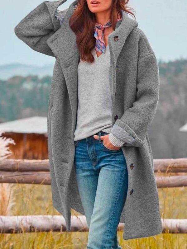 Vrouw draagt de lange grijze jas met capuchon open over een grijze trui, jeans en een kleurige sjaal. 2