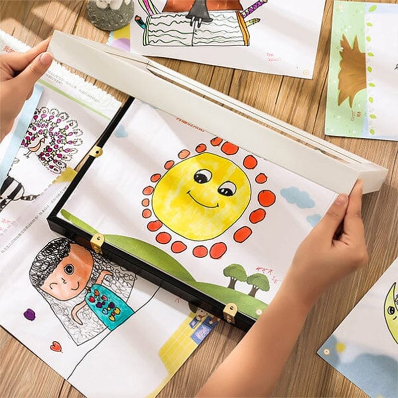 Kinderhanden openen een lijst boven een zonnetekening, omringd door andere kindertekeningen op een houten tafel. 12
