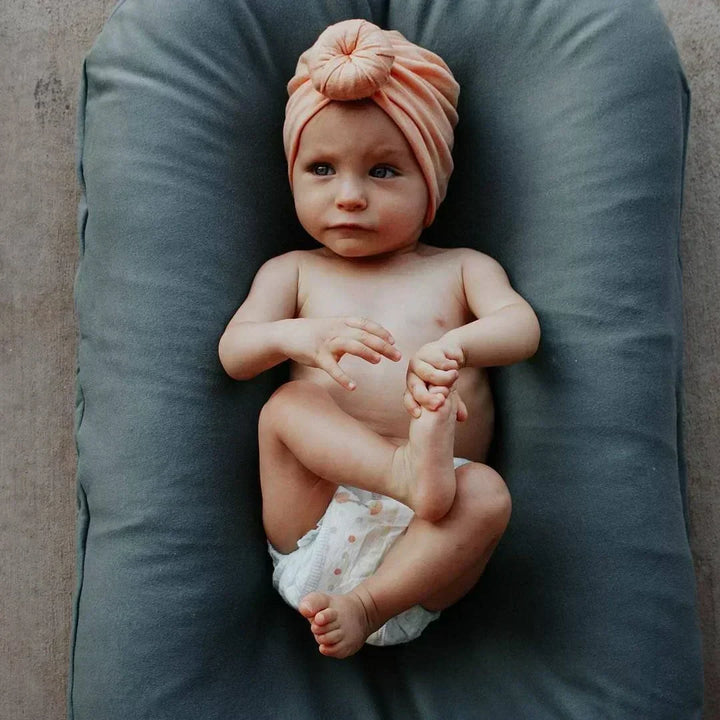 Baby met oranje tulband en luier ligt op een grijsblauw nestkussen en pakt zijn voetje vast 3