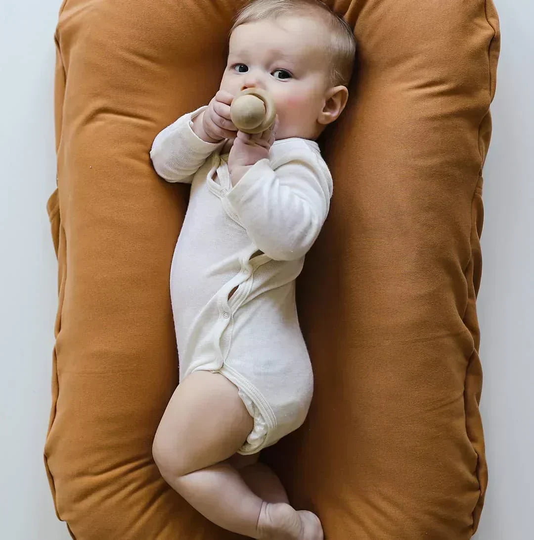 Baby in witte romper speelt met een houten bijtring in een oranjebruin nestje 4