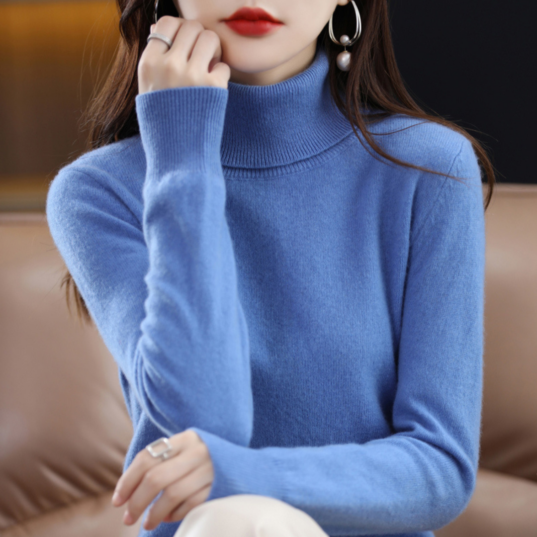 Blauwe coltrui gedragen door een model dat haar hand tegen haar kin houdt, zittend op een bruine bank. 3