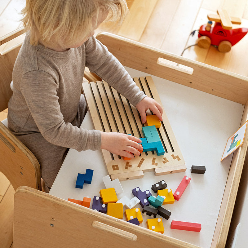 Kind zit aan een houten kindertafel en legt blokken op het bord, met losse blokken verspreid op het tafelblad. 8