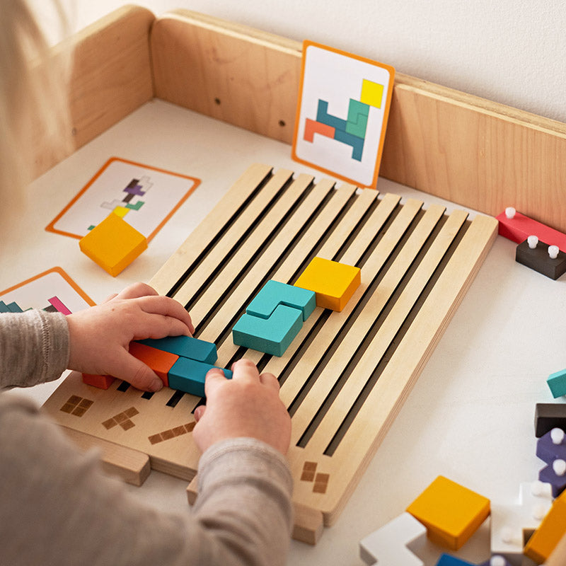 Kind legt met beide handen gekleurde blokken in de sleuven van het houten bord, met opdrachtkaarten op de achtergrond. 3
