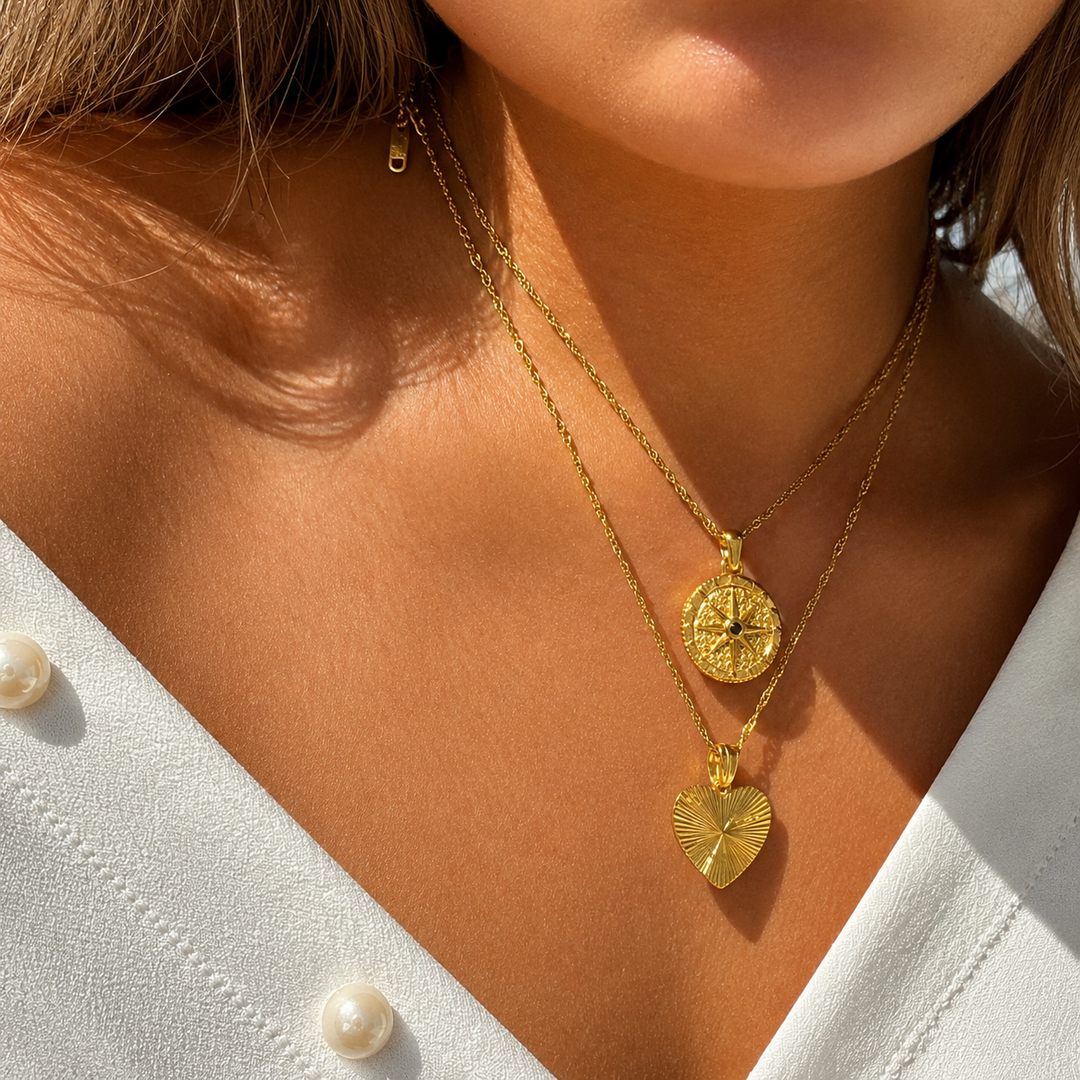 Close-up van een vrouw die de kompashanger draagt, samen met een tweede gouden ketting met hartvormige hanger, bij een witte blouse. 3