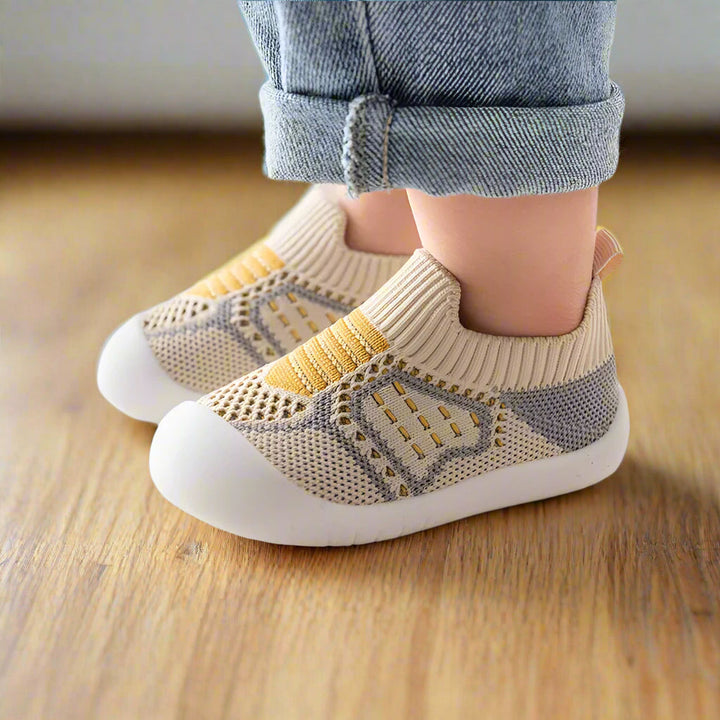 Zijaanzicht van kindervoeten in beige gebreide schoentjes met gele streepdetails en witte zool op een houten vloer. 9
