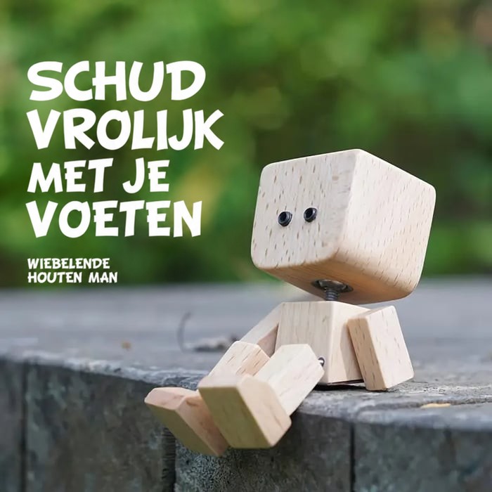 Houten figuurtje met zwarte oogjes zit met bungelende benen op een stenen muurtje voor een groene, wazige achtergrond. 13