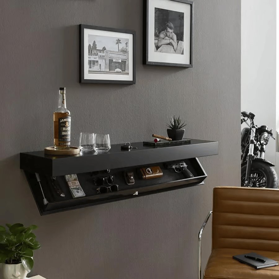 Zwarte muurplank aan een grijze muur in een woonkamer, met open verborgen vak onder een fles, glazen en plantje, naast een leren stoel. 4