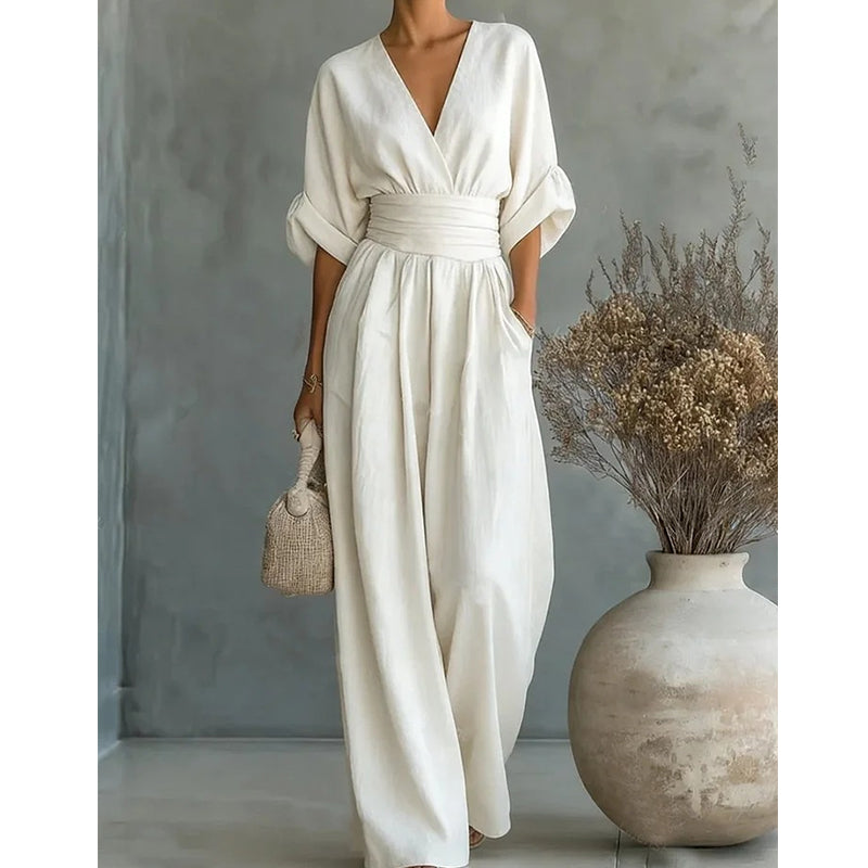 Crèmewitte jumpsuit op iets lichtere achtergrond, gedragen door een model met rieten tasje naast een vaas met droogbloemen. 6