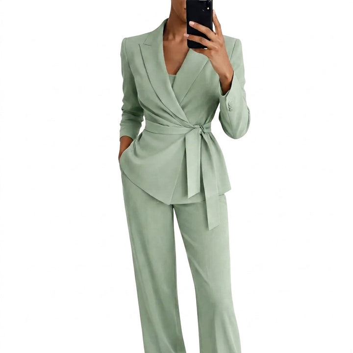 Vrouw in lichtgroen pak met overslagblazer, strikceintuur en wijde broek, die met een telefoon een spiegelfoto maakt. 1