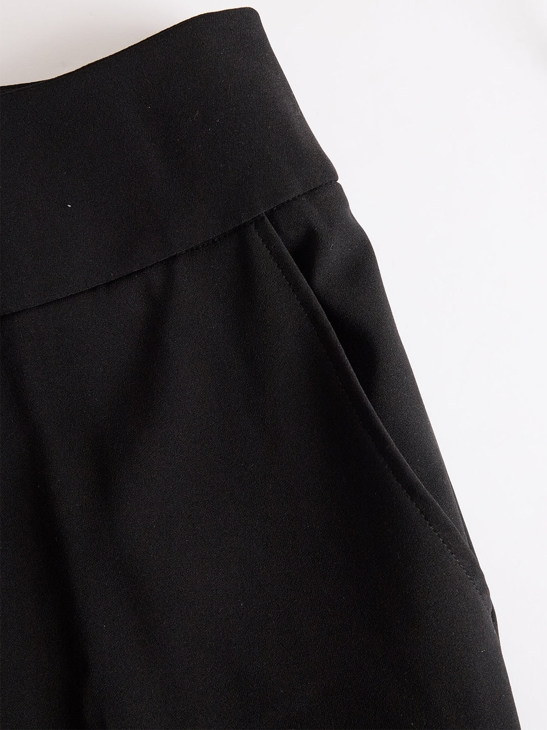 Detail van de zwarte broek met brede tailleband en steekzak, plat neergelegd op een witte achtergrond 5