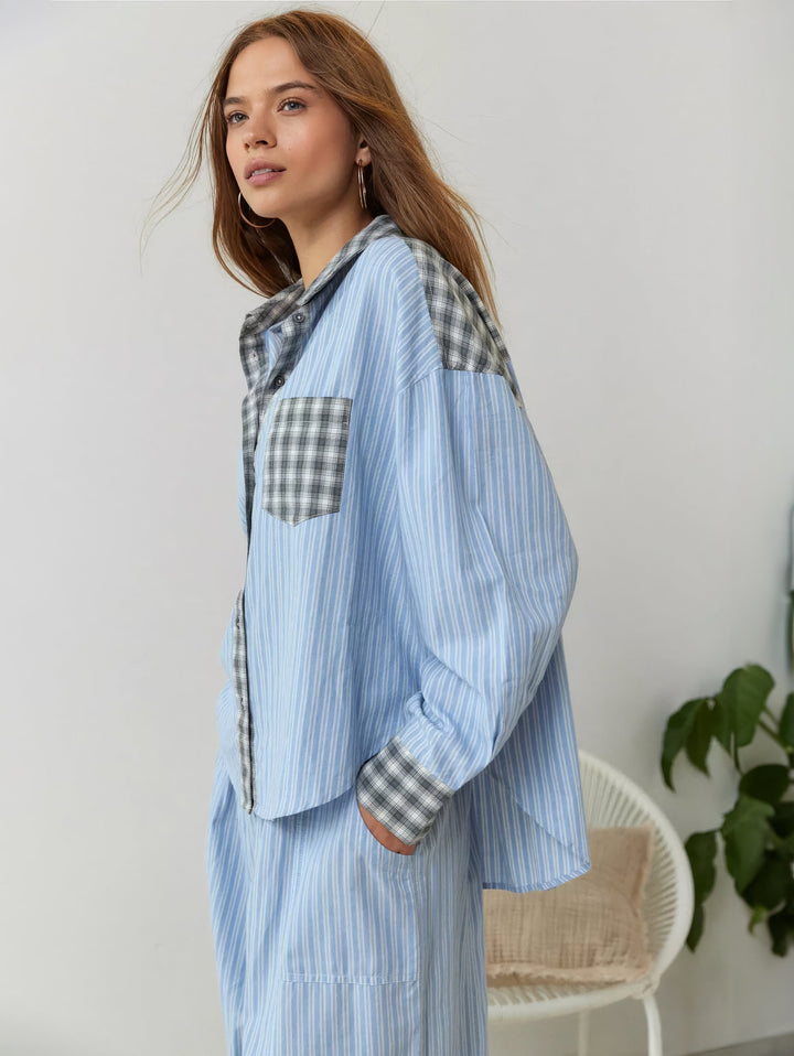 Zijaanzicht van een model in blauw gestreept overhemd met grijs geruite details en bijpassende broek, naast een wit rieten stoeltje. 3
