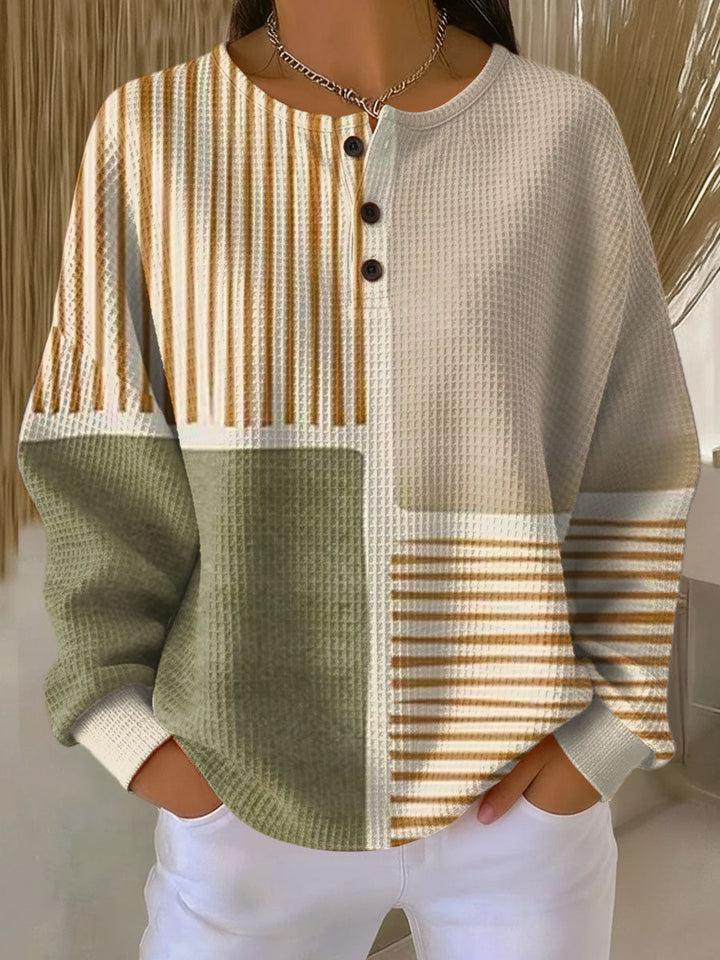 Wafeltrui met knoopjes aan de hals en kleurblokken in groen, beige en bruingeel gestreept, gedragen met witte broek. 1