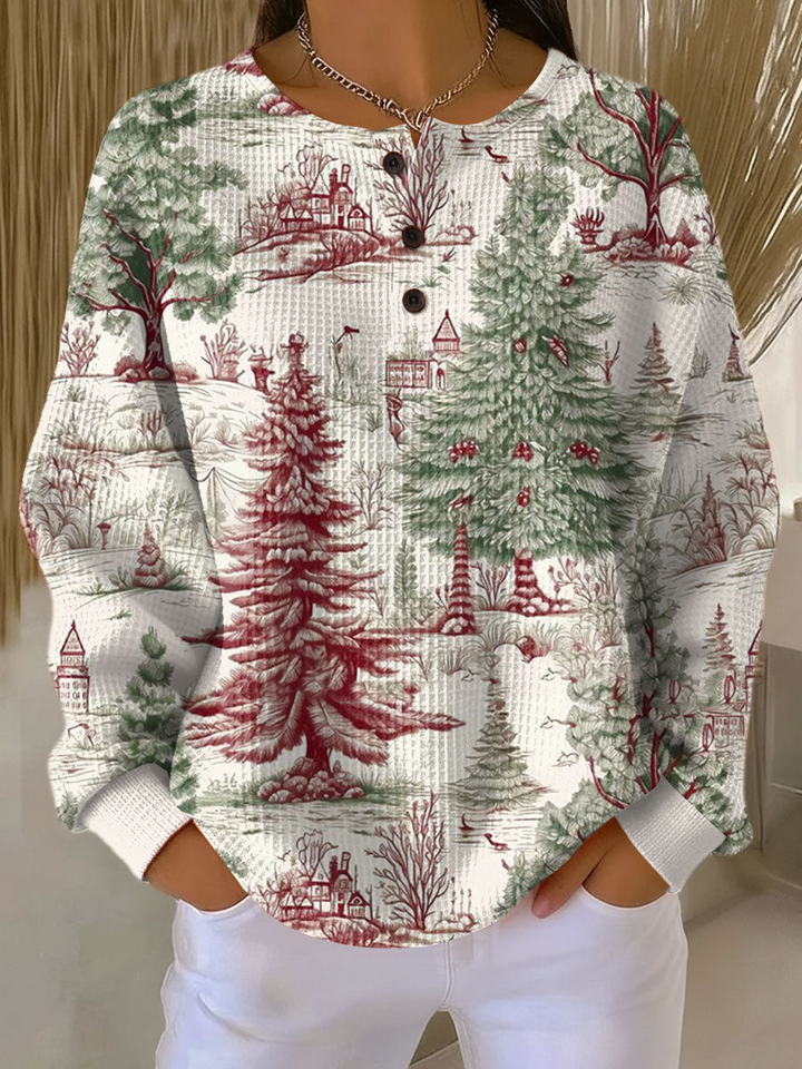Crèmewitte wafeltrui met knoopjes en een rood-groene winterprint van dennenbomen, huisjes en kale bomen, gedragen met witte broek. 22